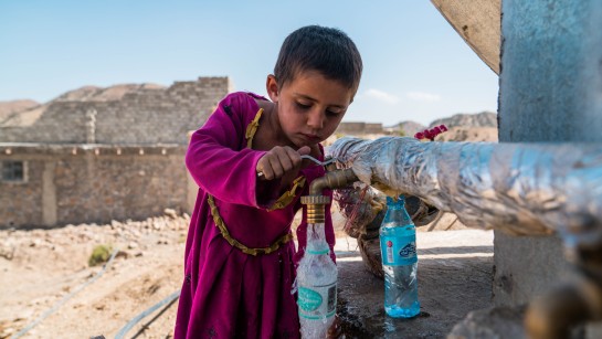 En pojke i Afghanistan fyller på vatten i sin flaska. Hans familj har fått tak över huvudet i en nybyggd bosättning som UNHCR byggt. Bostäöderna ger familjer ett bättre skydd under vintern. Varje hus är utrustat med traditionella vedeldade bukhari-kaminer för att hålla värmen, solpaneler som ger nödvändig belysning samt moderna latriner för bättre sanitet och hygien.