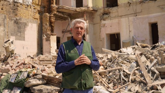 UNHCR:s chef Filippo Grandi står framför ett sönderbombat hus i Ukraina.